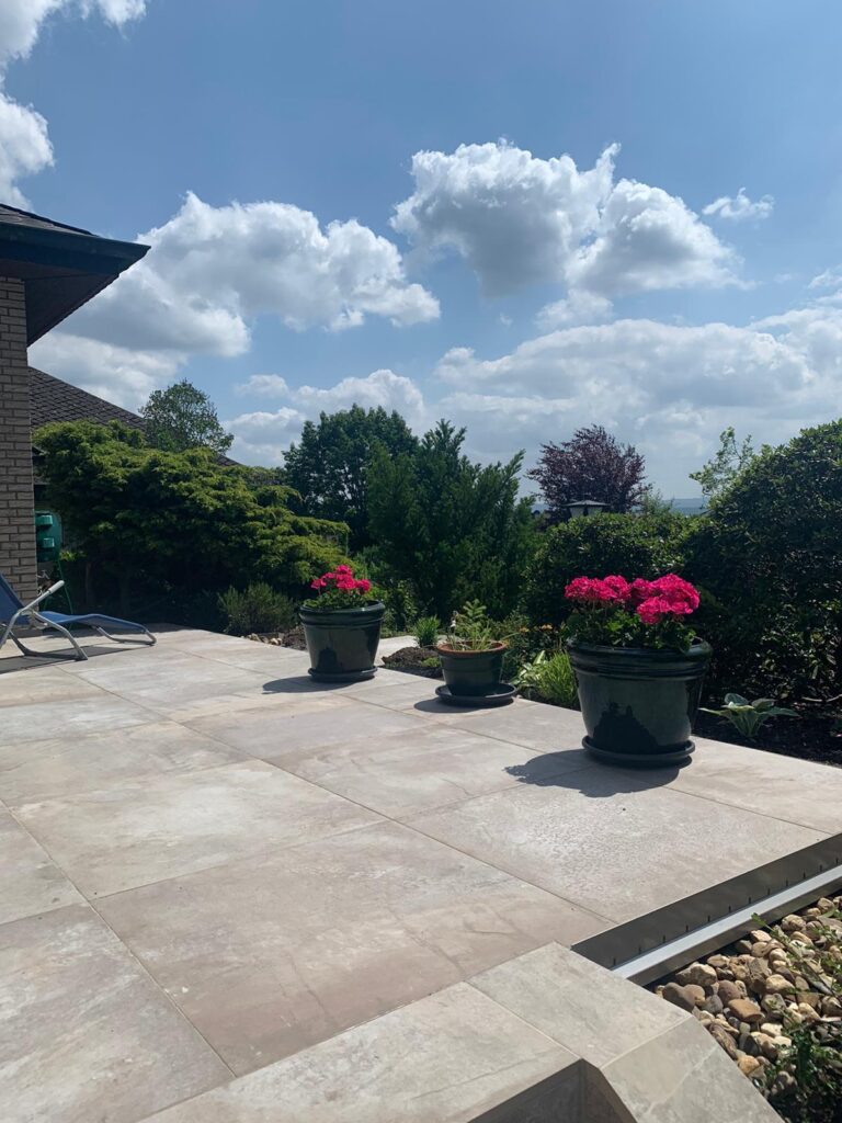 Terrasse mit Gartenanteil, Auf der Terrasse stehen Blumentöpfe mit Geranien. Die Terrasse wurde mit Terrassenfliesen in Steinoptik gefliest